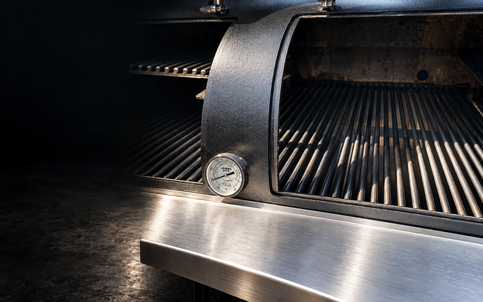 Jambo cook chamber open, showing heavy grates, a Jambo Pits temperature gauge and a stainless shelf