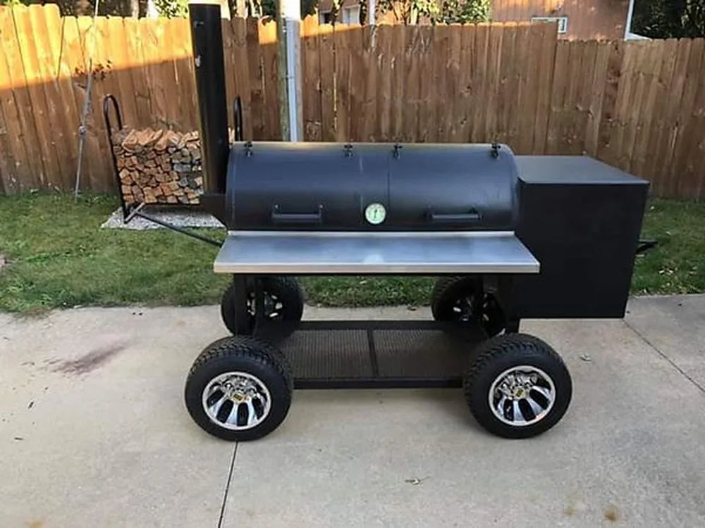 Black Jambo Baby J with stainless shelf on large custom wheels, beside a stack of firewood